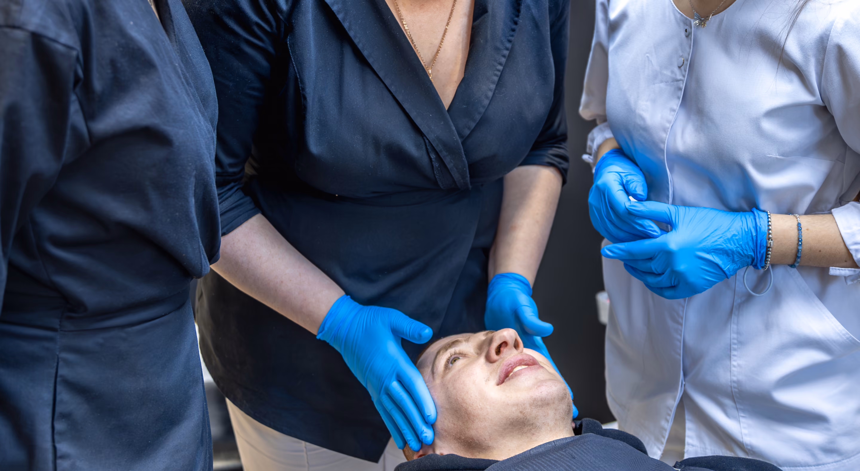 A man lying back while two people wearing blue gloves and dark clothing gently examine or massage his head.