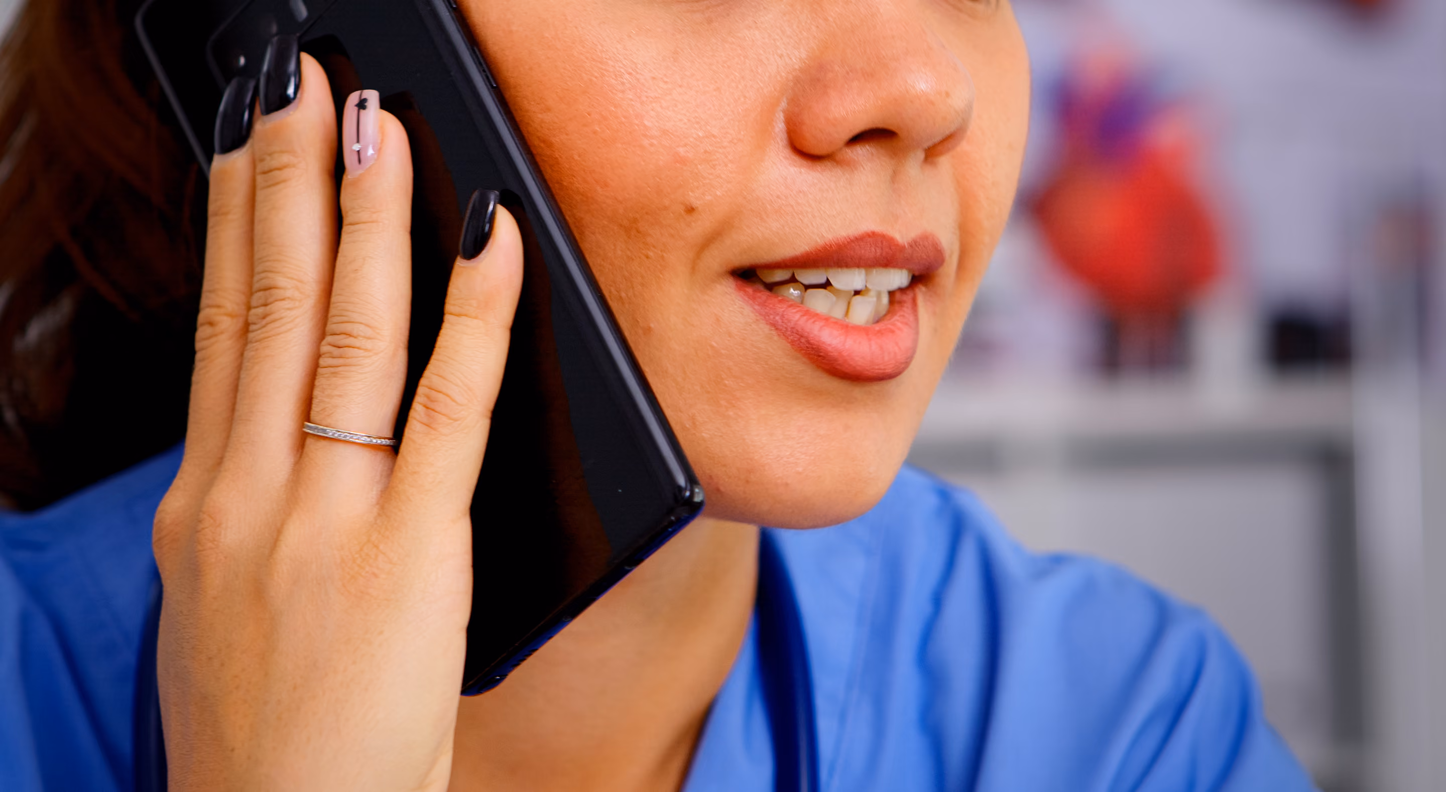 Close-up of a woman in blue scrubs holding a black smartphone to her ear, smiling while speaking.