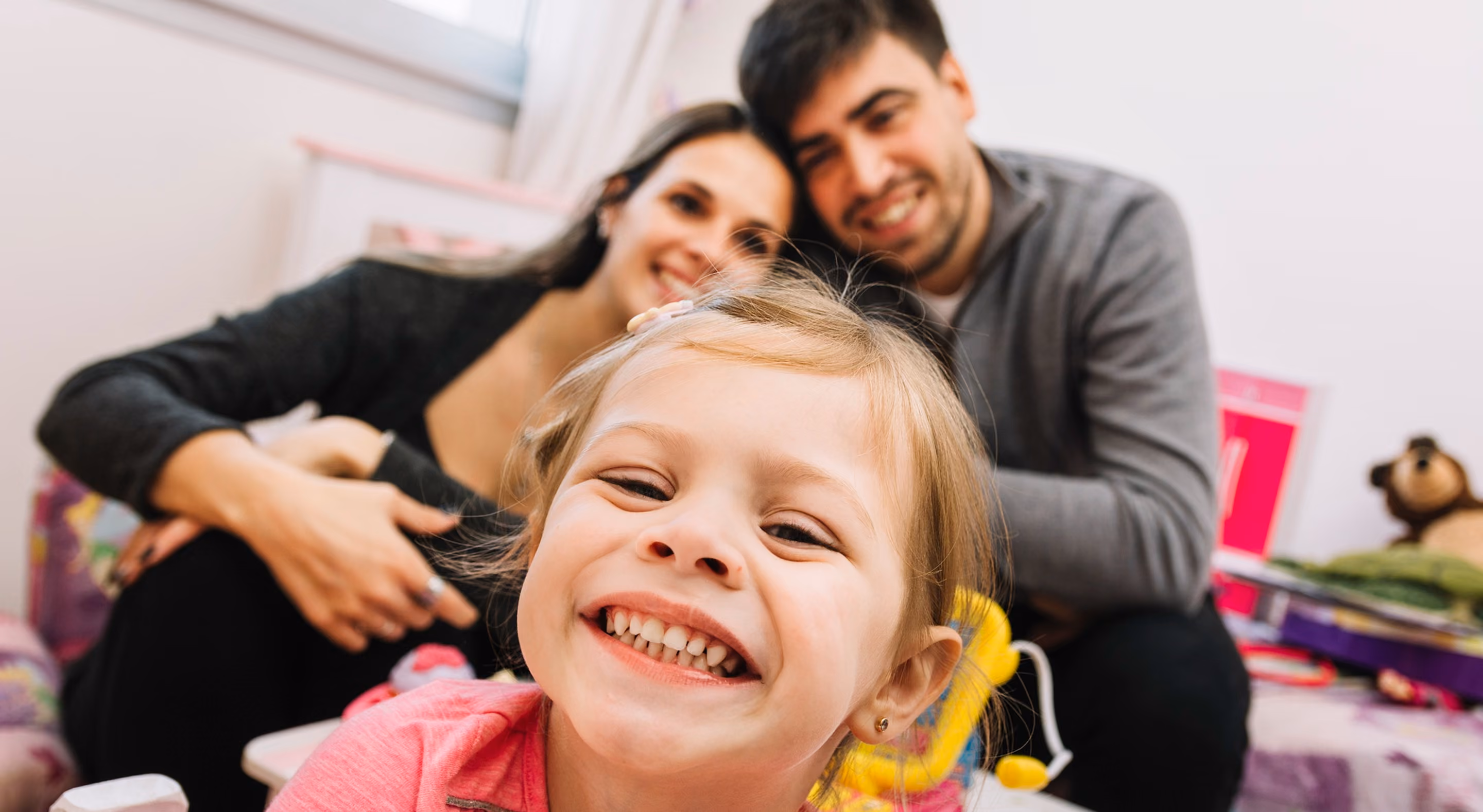 Close-up of a smiling young girl in the foreground with two adults, likely her parents, smiling in the background inside a room with toys.