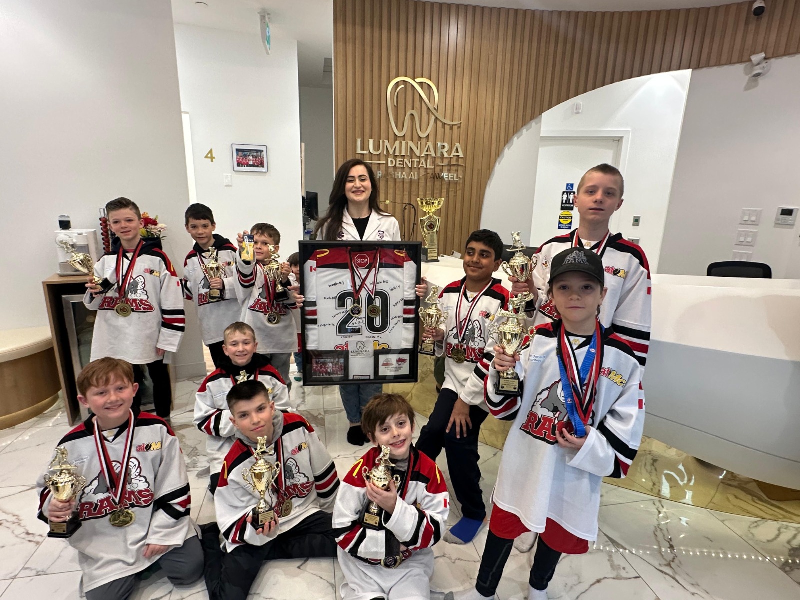 A group of young hockey players in matching white Rams jerseys with red and black details, holding trophies and medals, posing indoors in front of a wooden wall with a Luminara Dental sign and a woman in a white coat holding a framed hockey jersey signed by the team.