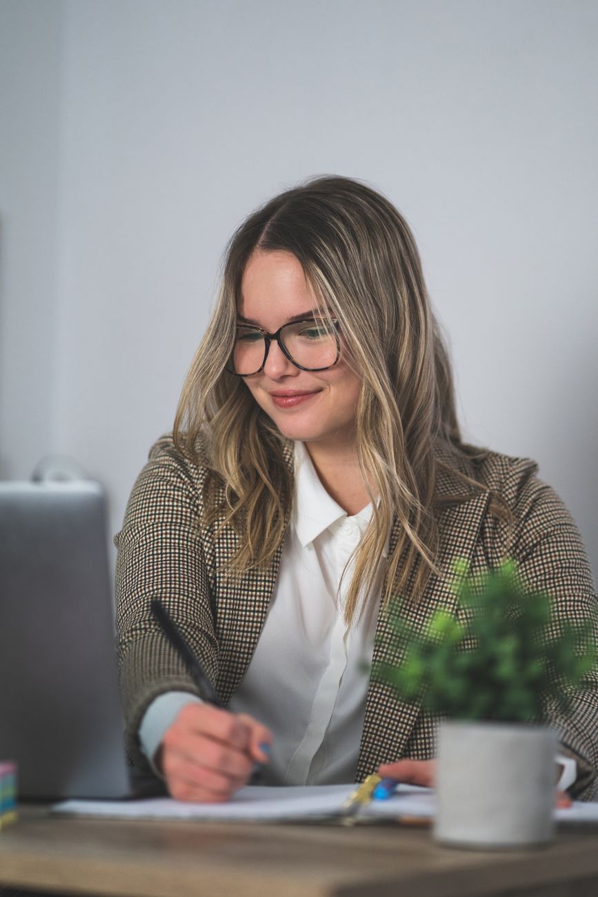Smiling woman in plaid blazer writing at desk with plant and laptop.