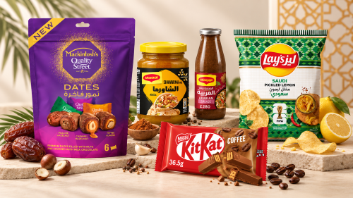 Assorted packaged foods displayed on a beige surface: a purple packet of Mackintosh's Quality Street dates with hazelnuts, a jar of Maggi Shawarma seasoning, a bottle of Maggi Arabic sauce, a green and white bag of Lay's Saudi Pickled Lemon flavored chips, and a Nestlé KitKat Coffee chocolate bar with coffee beans scattered around.