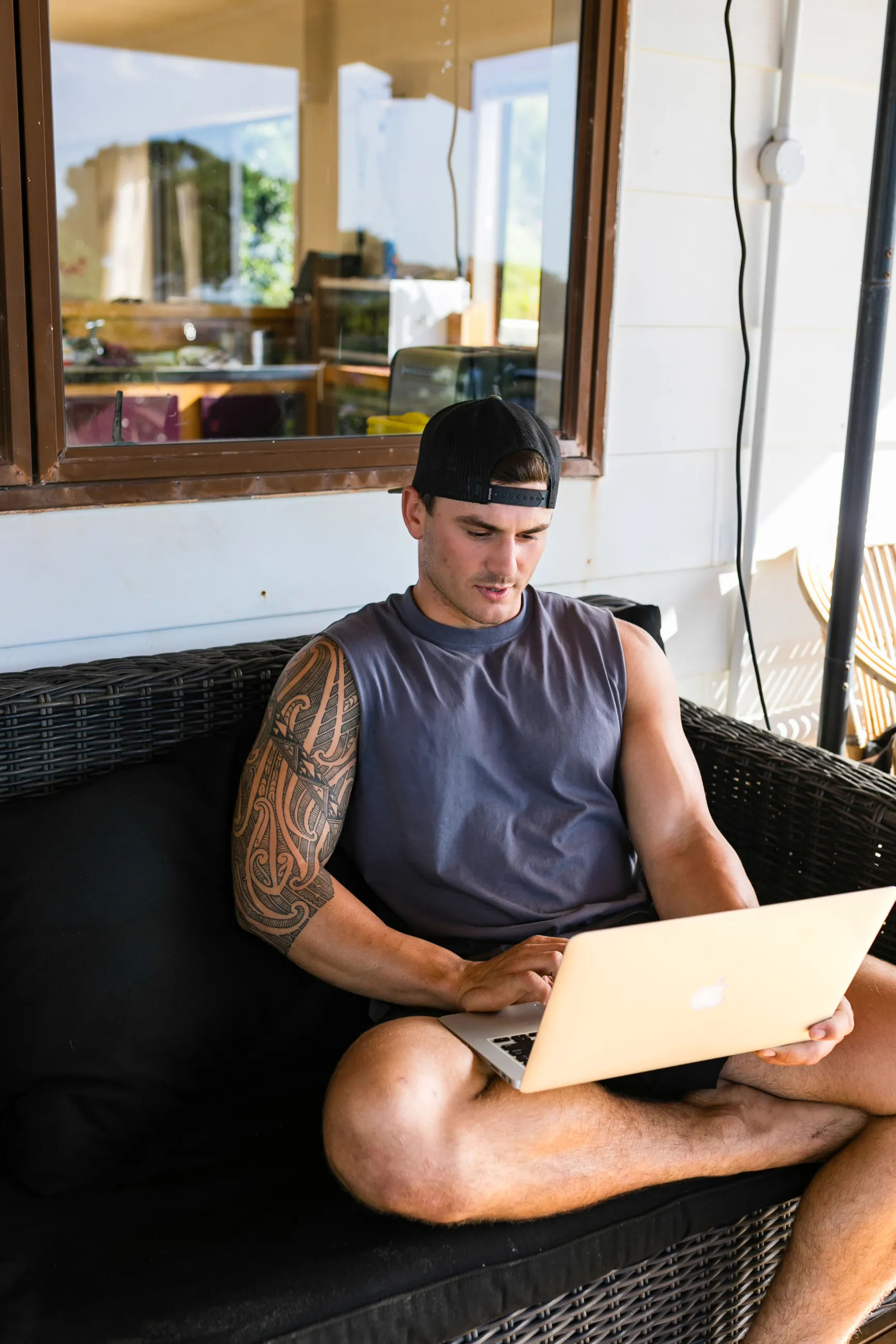 Man with a tattoo sleeve on his left arm wearing a backward cap and sleeveless shirt, sitting on a wicker sofa using a laptop.
