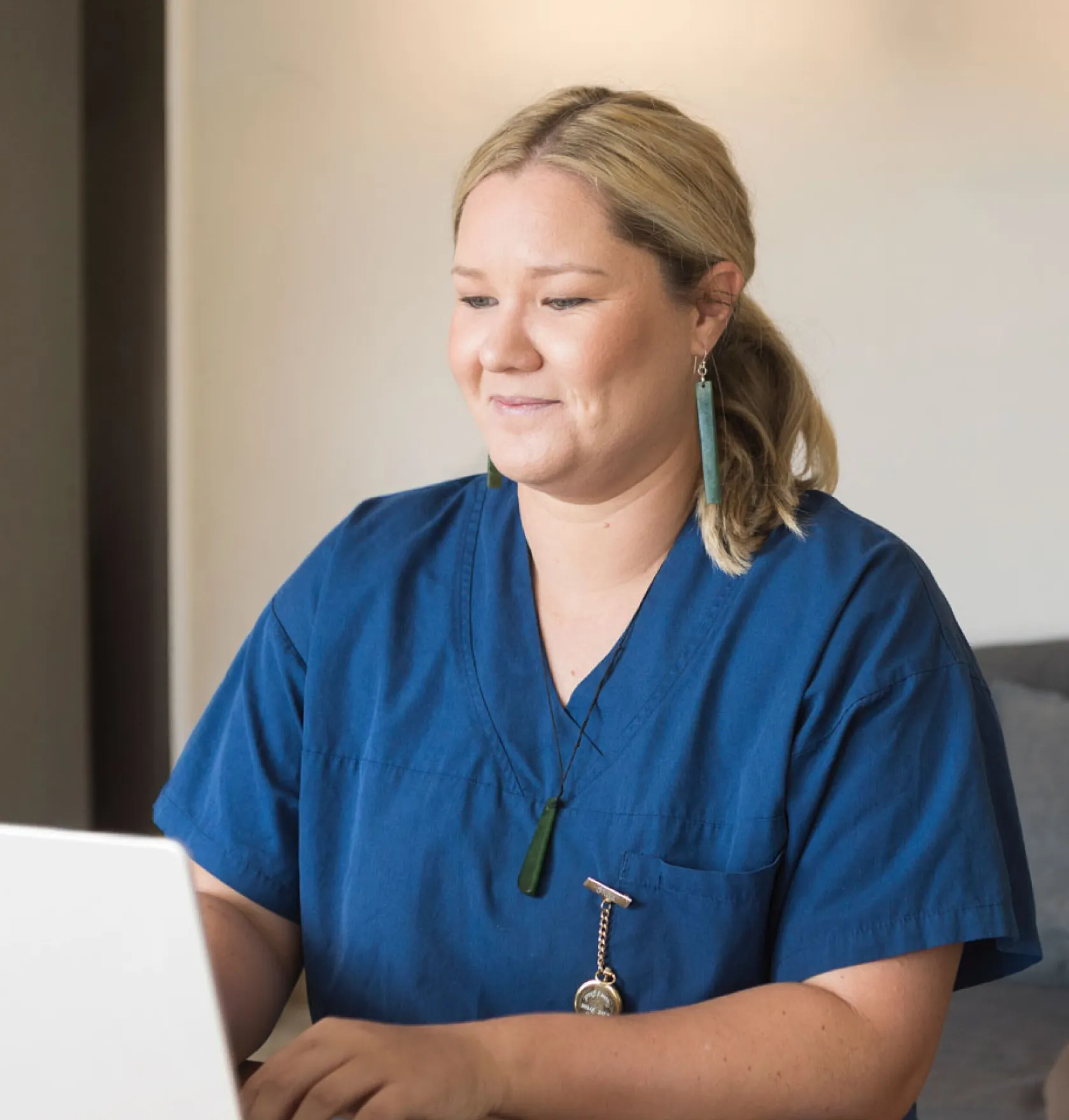 Smiling woman wearing blue medical scrubs working on a laptop indoors.