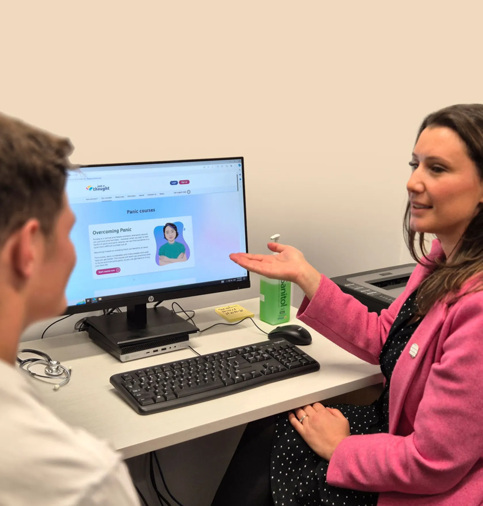 A woman in a pink jacket discusses panic courses displayed on a computer screen with a man in a white shirt.