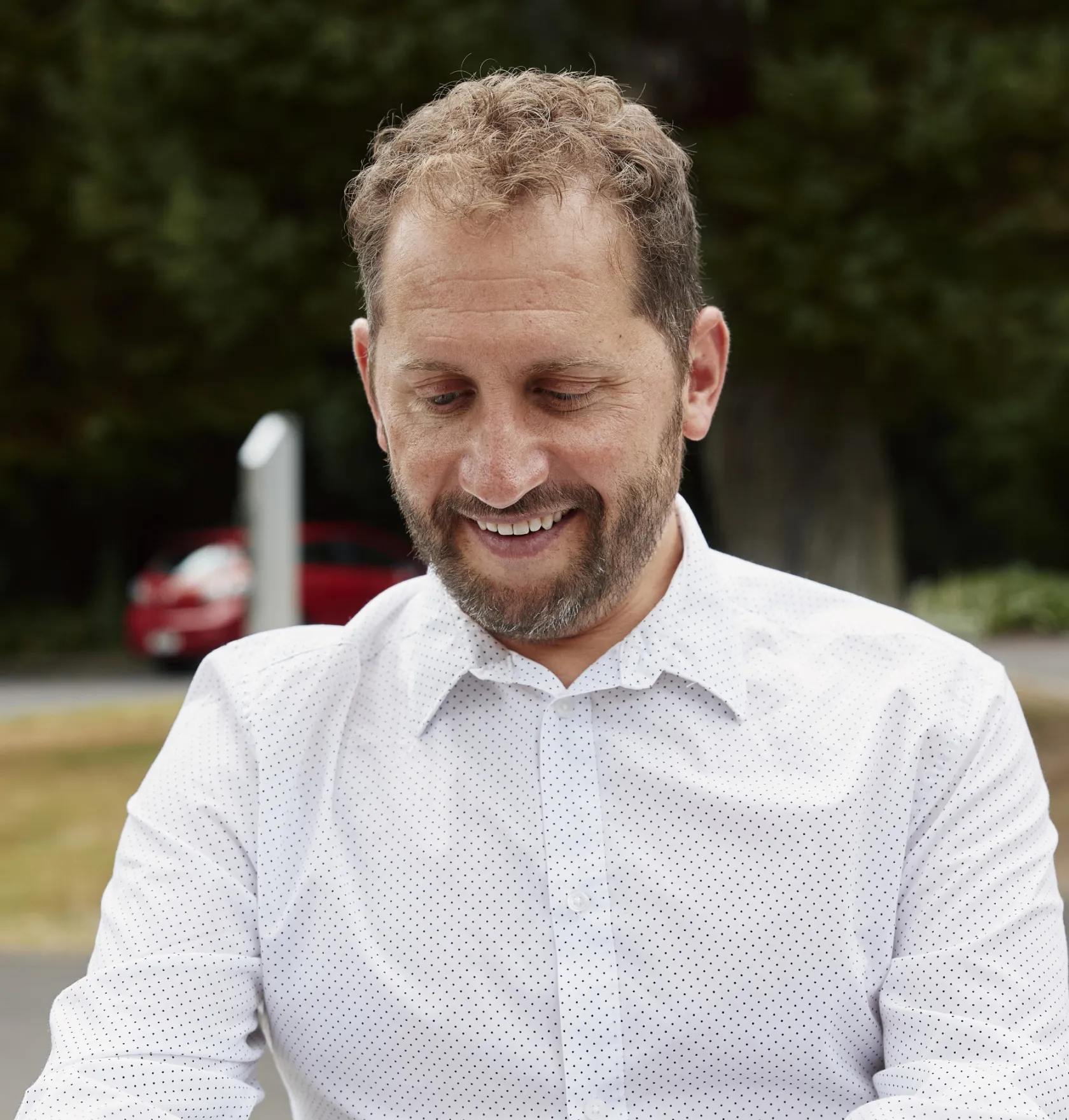 Smiling man with short curly hair and beard wearing a white shirt with small black dots, outdoors with greenery and a red car in the background.