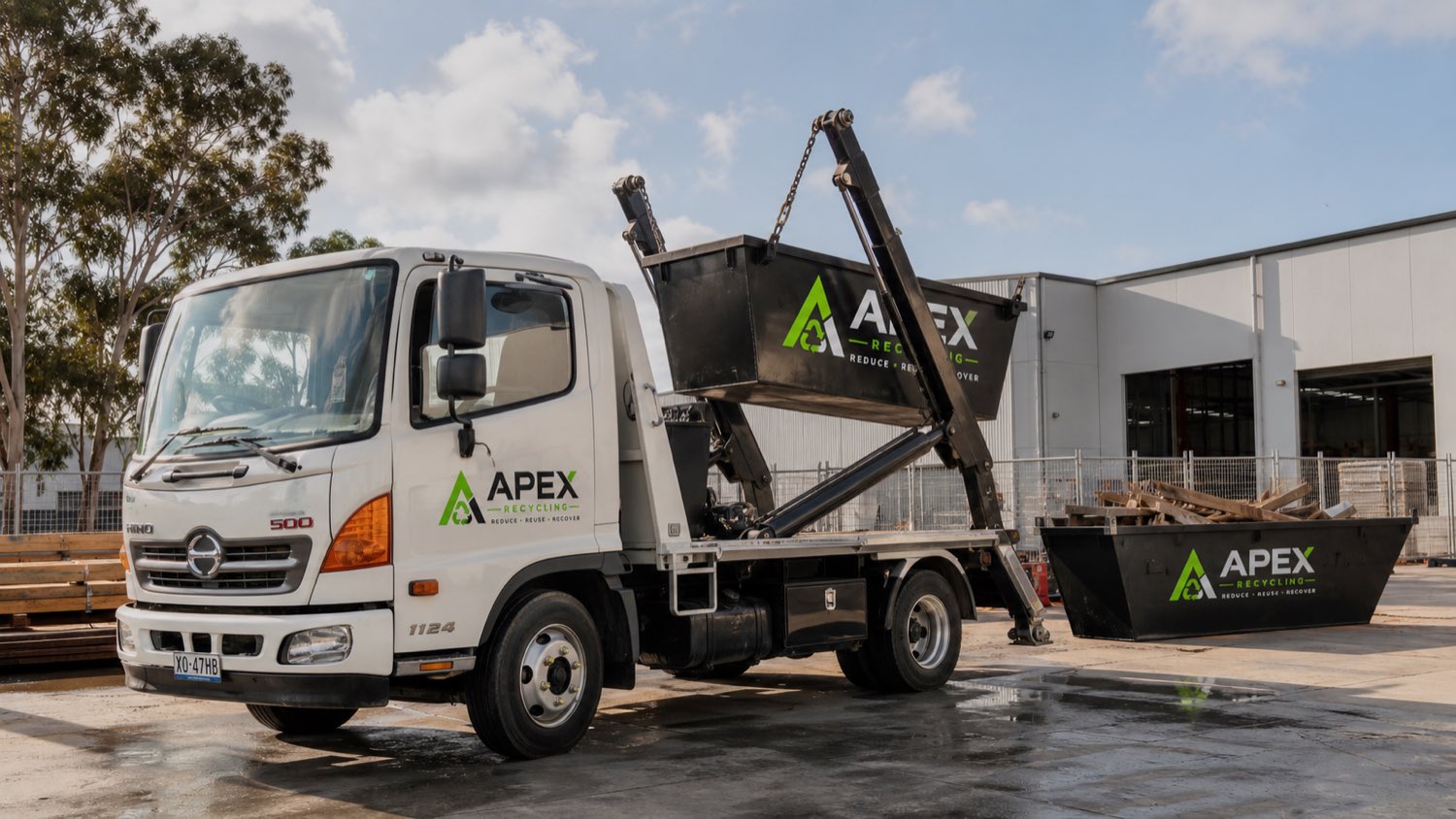 An APEX Recycling skip truck lifting a skip bin onto the tray, with a second bin alongside