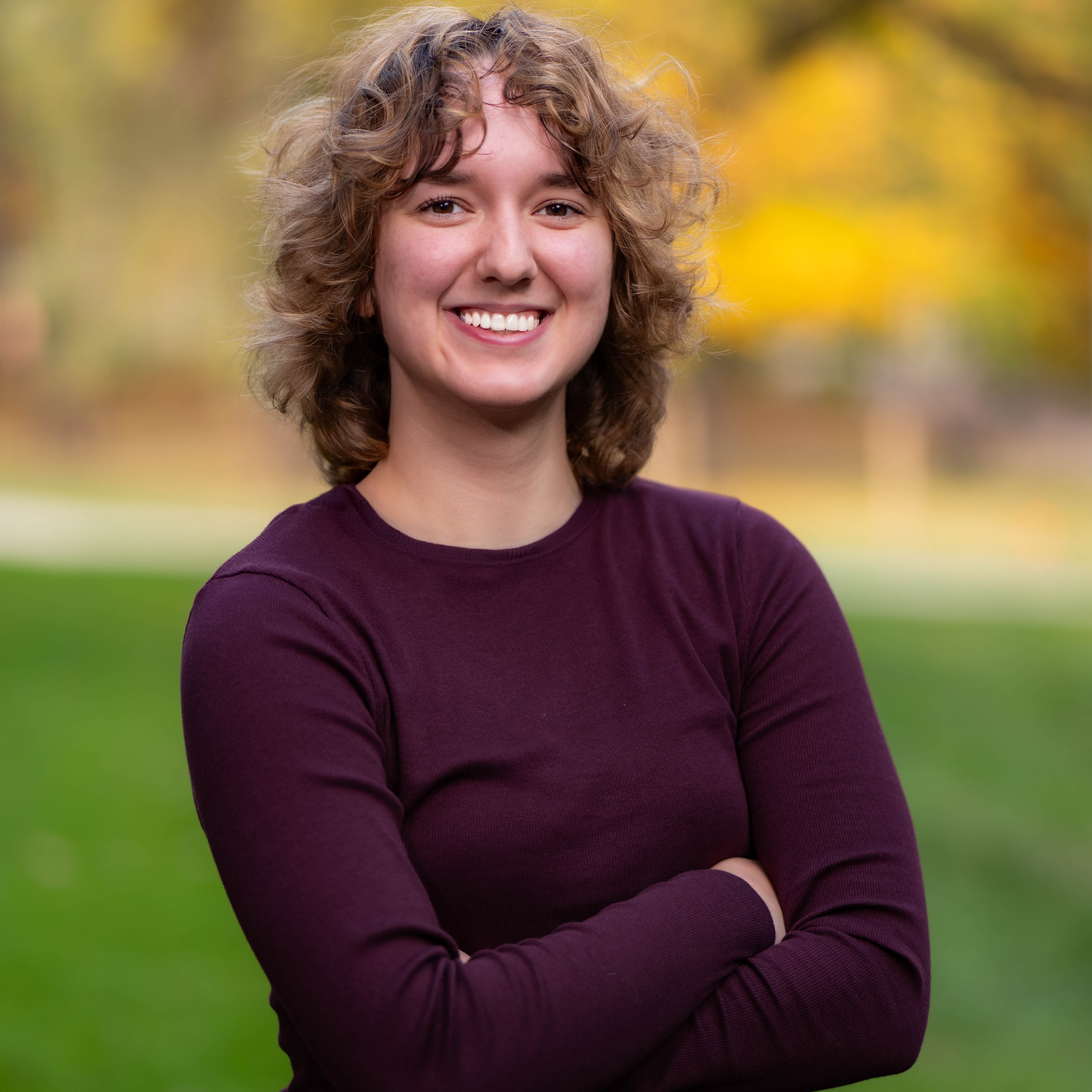 adria has mousy brown shoulder-length wavy hair, brown eyes, and is smiling. she is crossing her arms. she is wearing a redish-purple long sleeve shirt. the background is a fall nature scene with a blur effect.