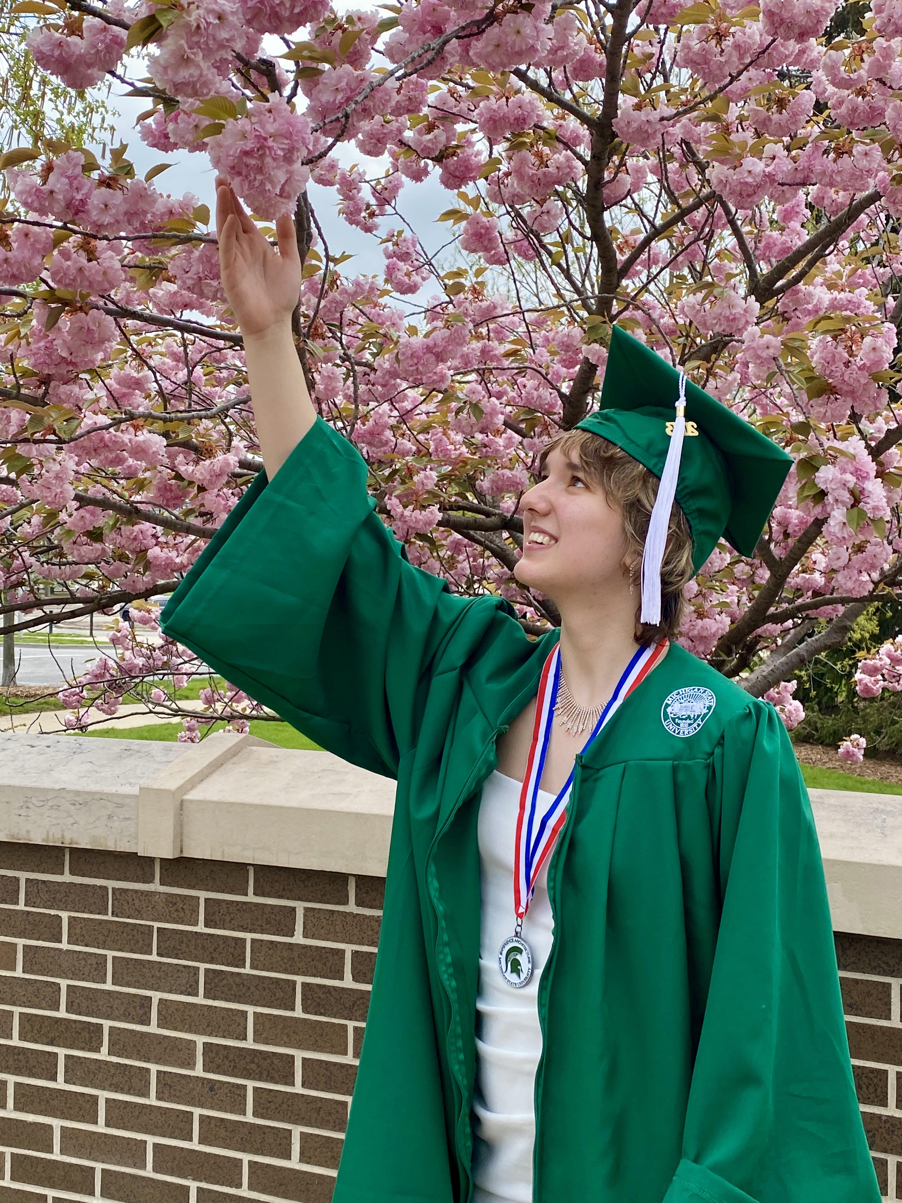 adria is wearing hergreen msu gown with a white dress. she as a medal around her neck with an msu logo medal with red, white, and blue stripes on the fabric of the medal. she looking up, holding her right arm up, holding a flower of the cherry blossom tree behind her.