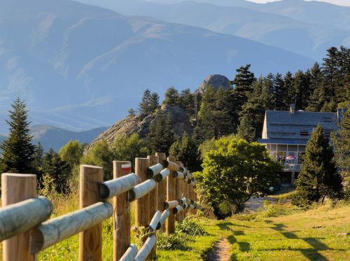 Wooden fence leading towards a house surrounded by trees on a grassy mountain slope, with layered blue mountains visible in the background.