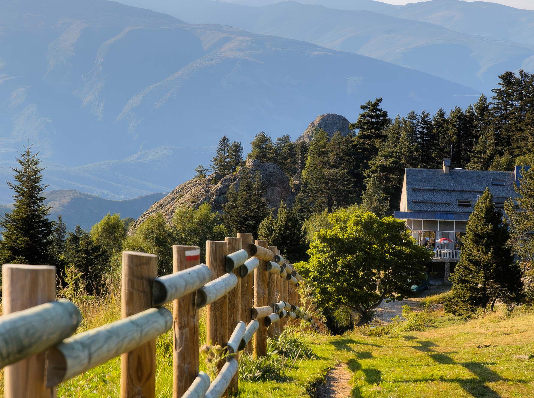 Wooden fence leading towards a house surrounded by trees on a grassy mountain slope, with layered blue mountains visible in the background.