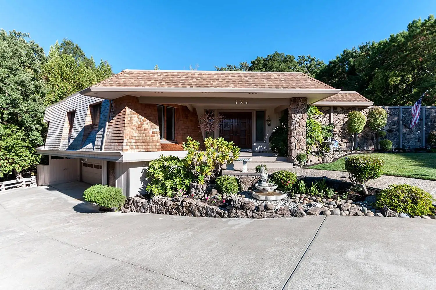 Mid-century modern home exterior featuring a concrete slab driveway, stone accent walls, wood shingle siding, and front yard landscaping.