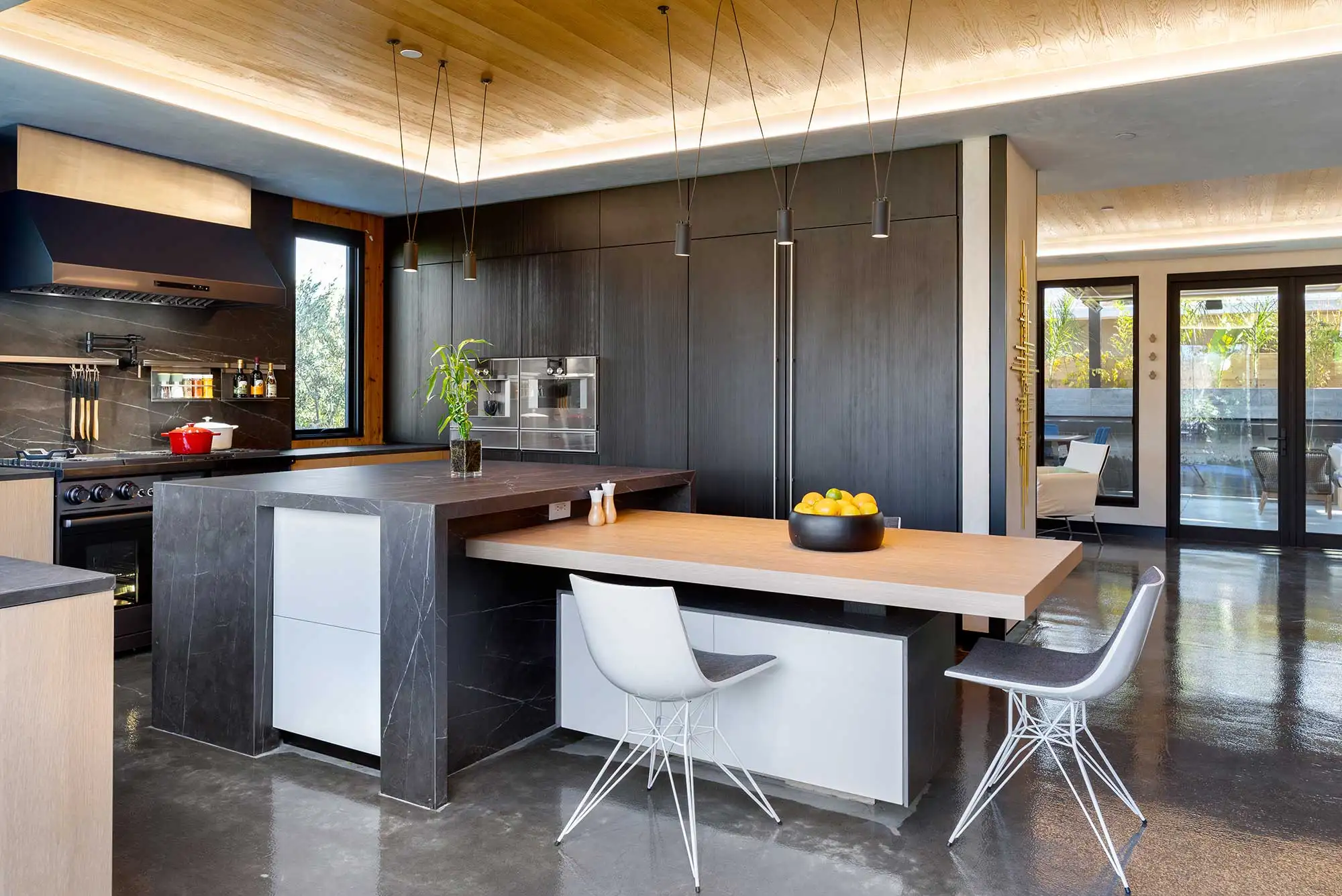 Custom luxury kitchen design following a pre-construction planning process, featuring dark slab island countertops, integrated wood dining extension, dark cabinetry, and polished concrete floors.
