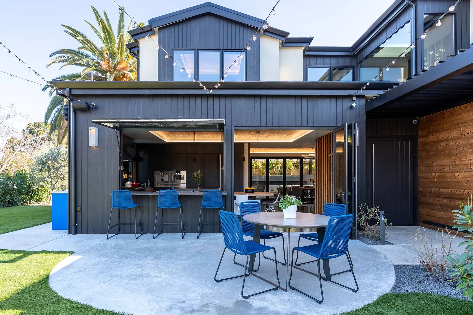 Modern two-story custom home back exterior with dark vertical siding, pass-through outdoor kitchen bar, folding glass doors, and concrete patio layout.