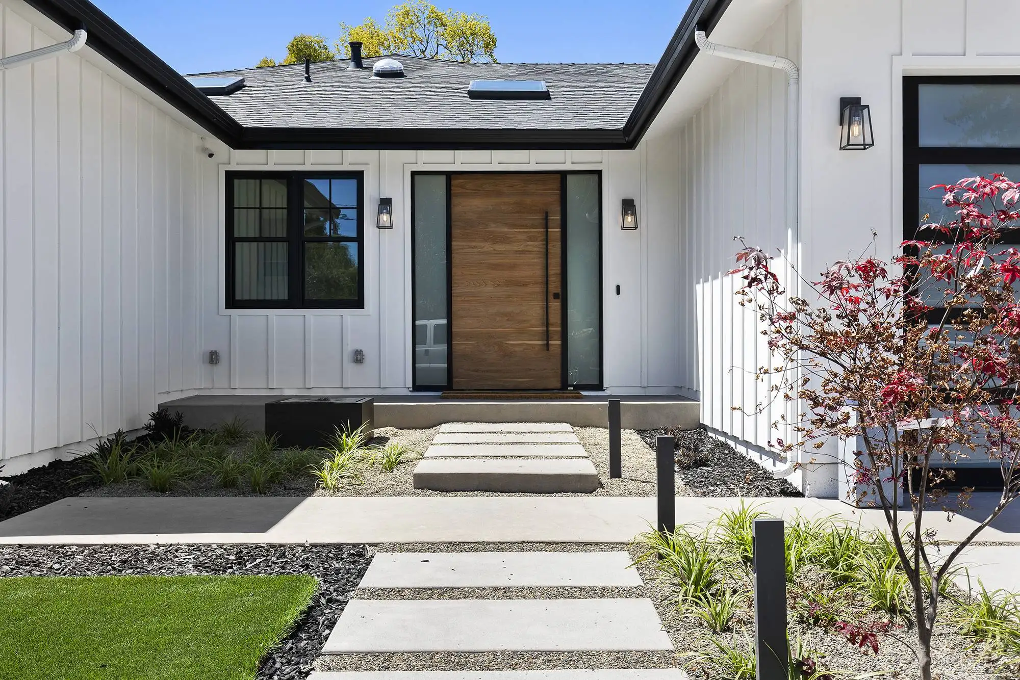 Modern custom farmhouse home entryway in San Carlos featuring white board and batten siding, a natural wood front door, concrete paver walkway, and contemporary landscaping.