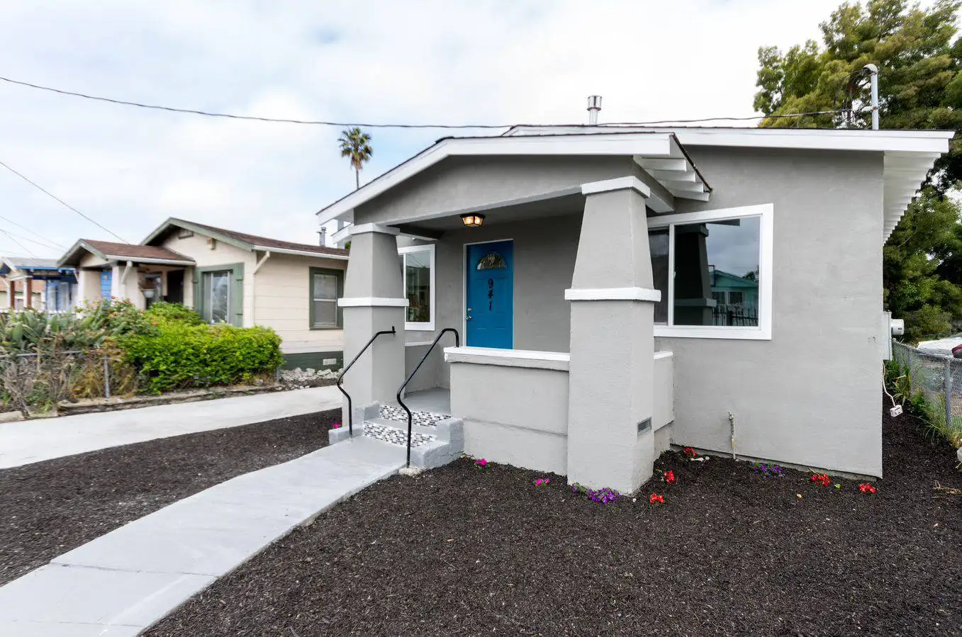 Remodeled bungalow home exterior featuring gray stucco siding, a blue front door, covered porch, and updated landscaping for residential home construction.