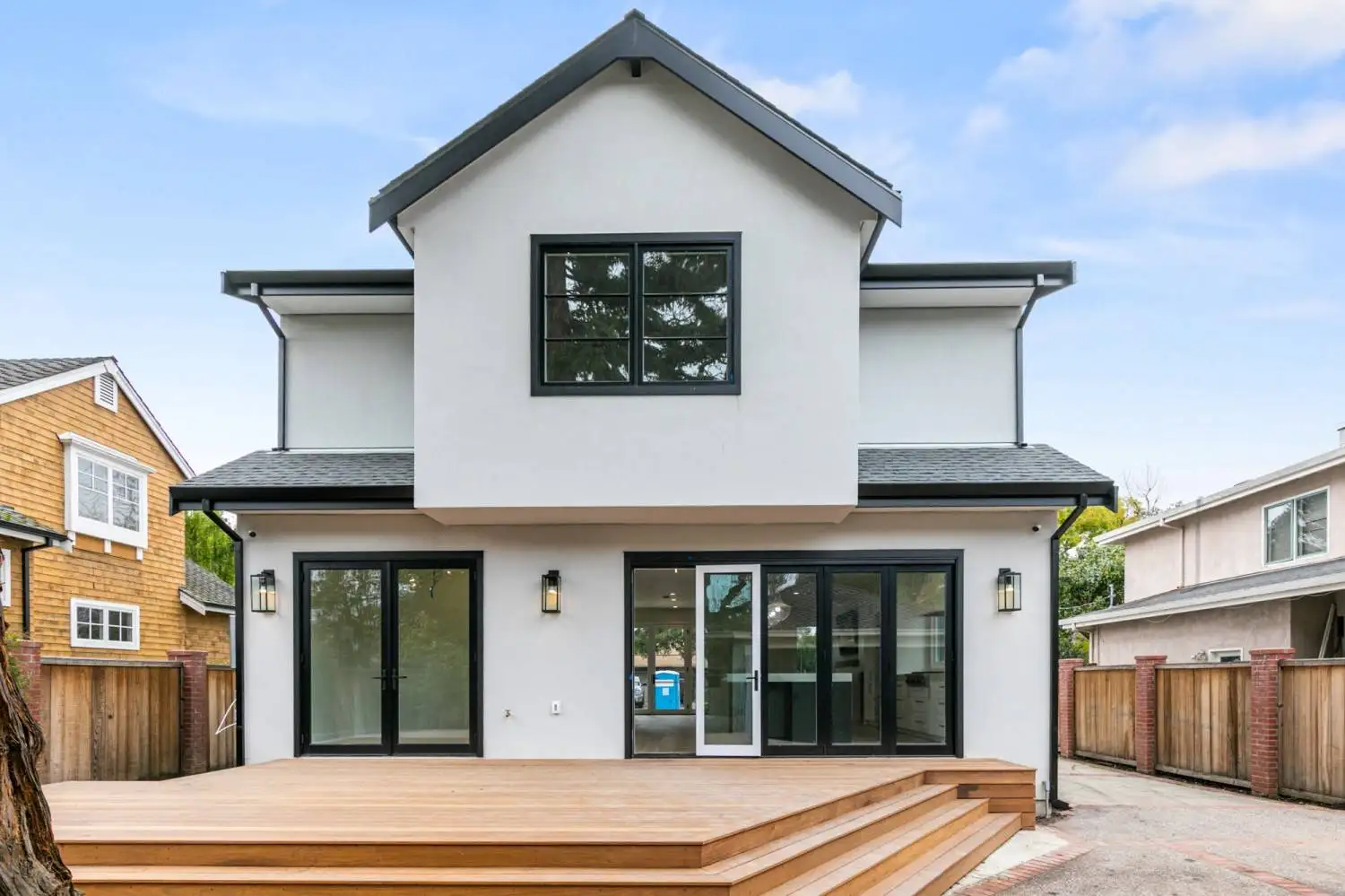 Rear exterior of a two-story white stucco home with black-framed windows, folding glass doors, and a wood deck