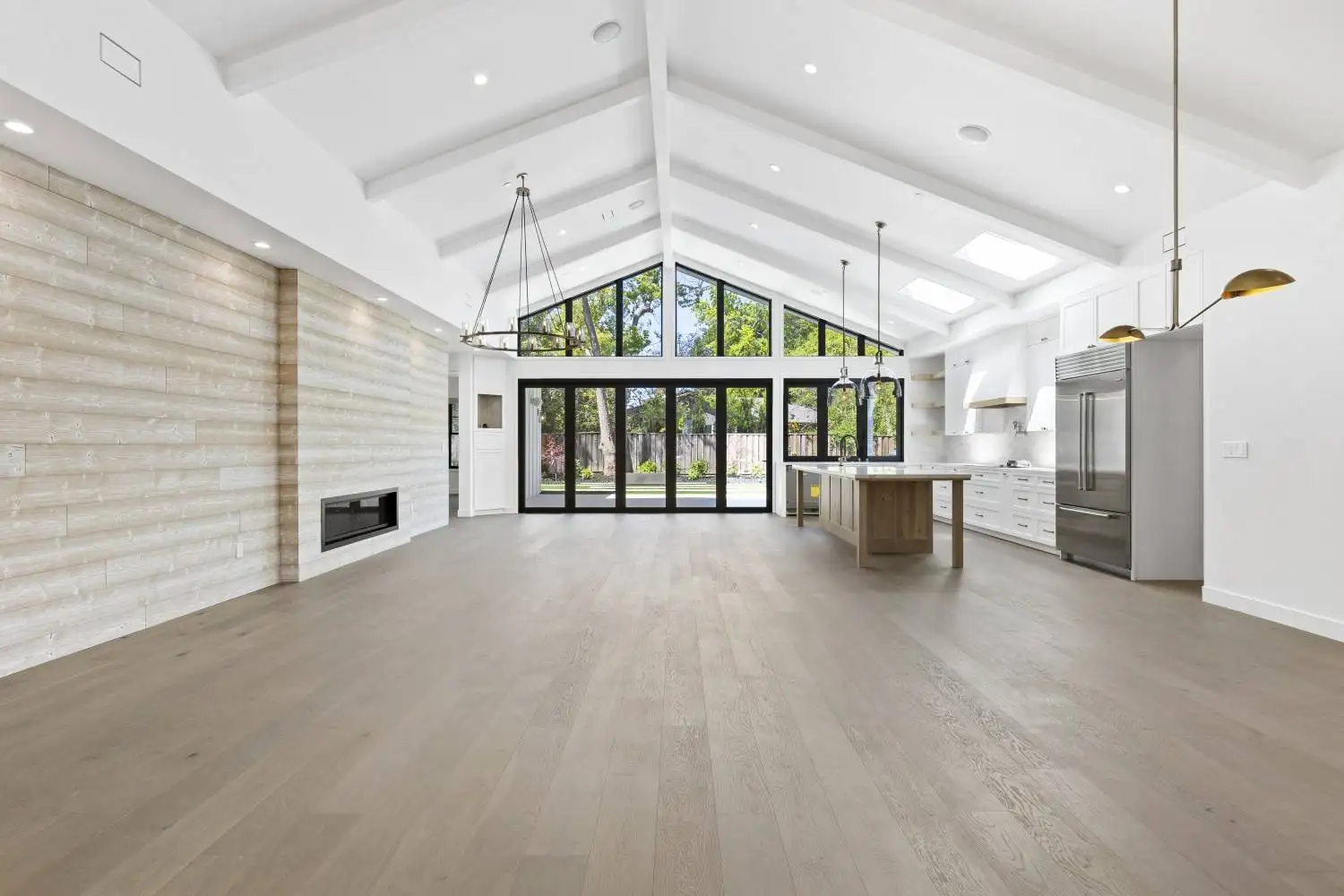 Open living and kitchen area under a vaulted beamed ceiling, with a wood accent wall, fireplace, and a large center island
