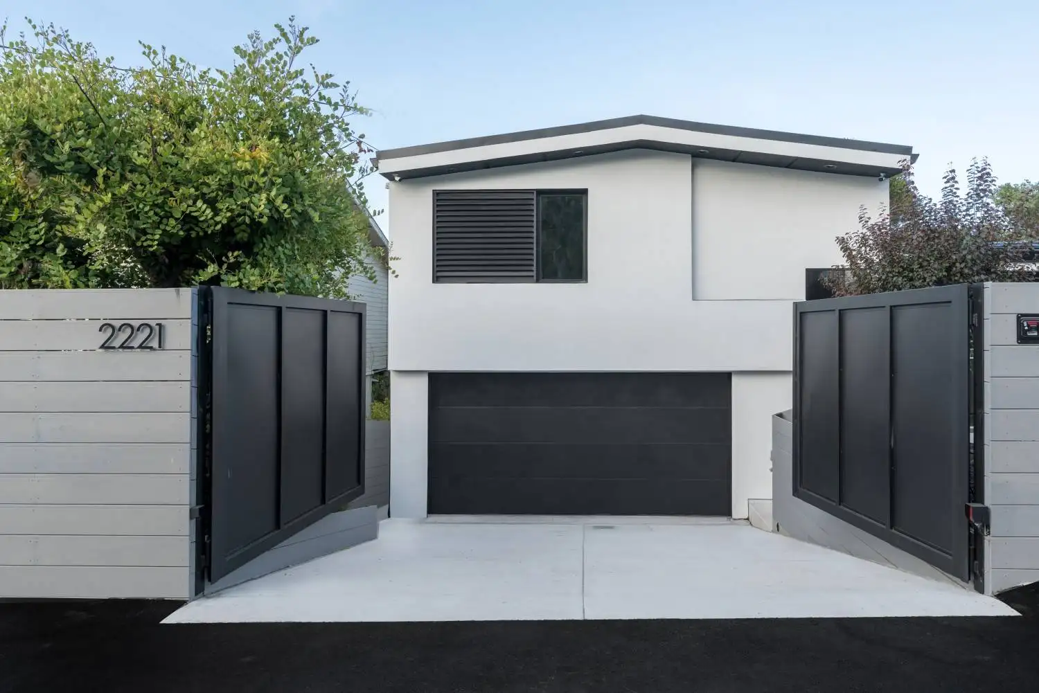 Modern home exterior behind an open dark metal driveway gate, with white stucco siding and a dark garage door