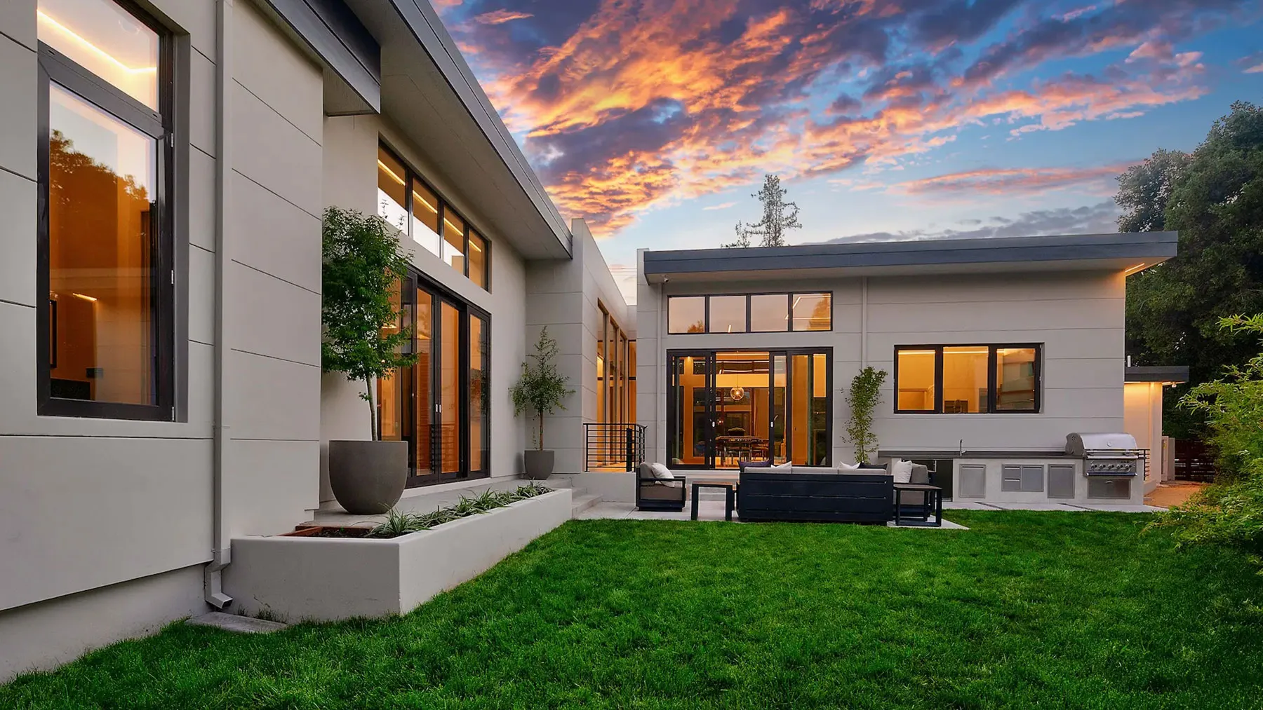 Modern luxury custom precision-built home exterior at sunset featuring floor-to-ceiling windows, an outdoor patio, built-in kitchen, and manicured green lawn.