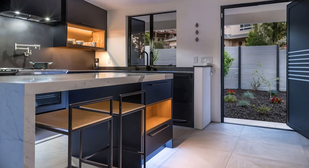 Modern kitchen with matte black cabinetry, marble countertop, wood shelving accents, and a door opening to a landscaped side yard