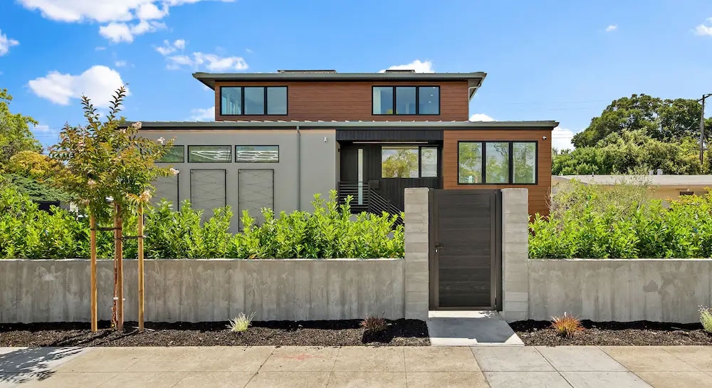 Modern two-story home exterior with wood and stucco siding, a low concrete perimeter wall, wood gate, and drought-tolerant landscaping