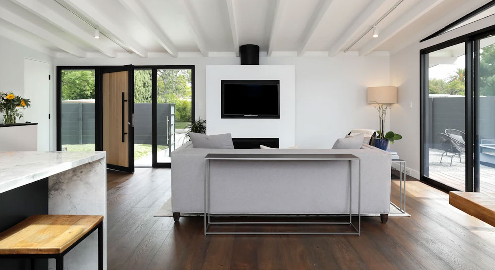 Open-concept living room with exposed wood-beam ceiling, wall-mounted TV above a fireplace, and sliding glass doors opening to a patio