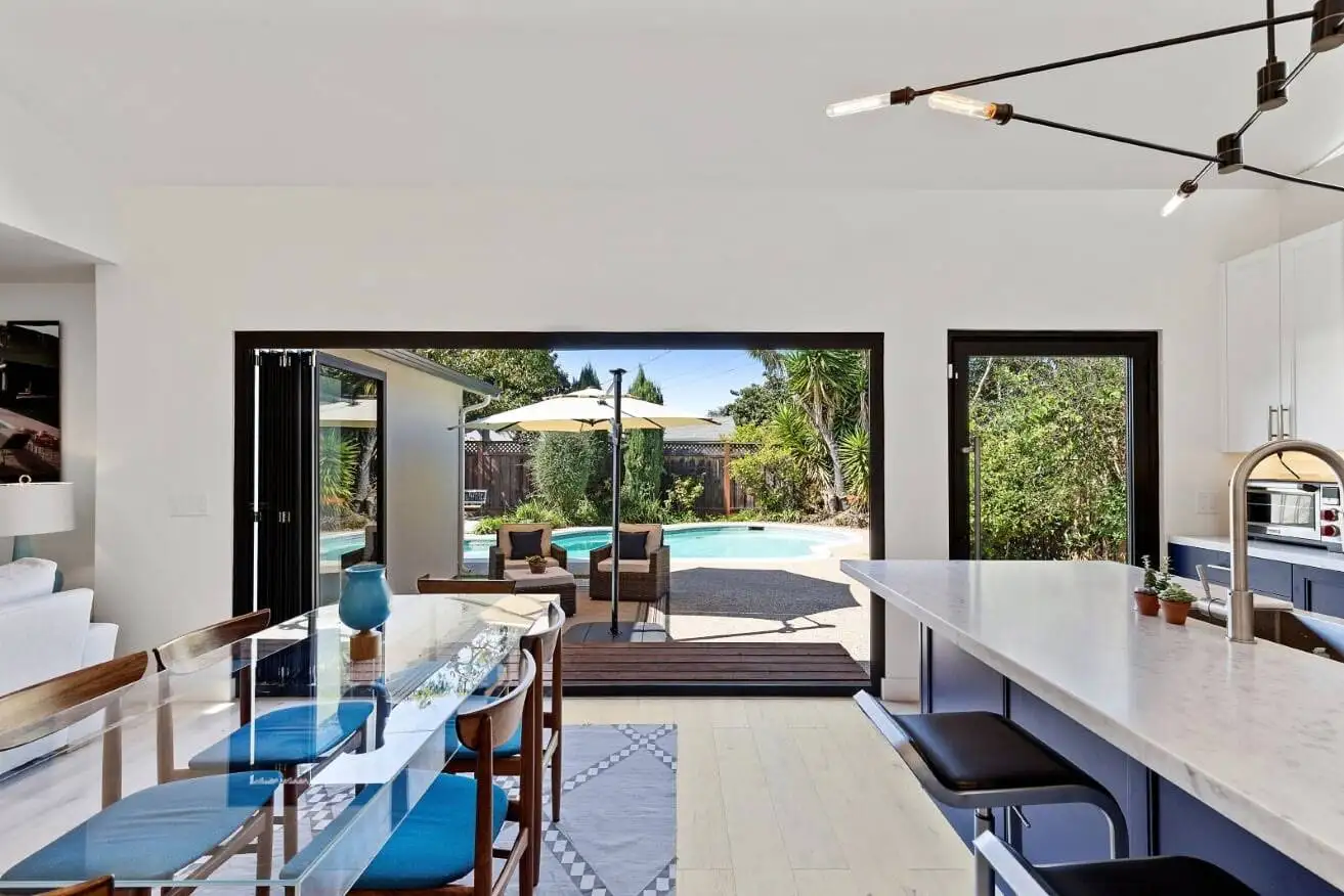 Open-concept kitchen and dining area with bifold glass doors opening to a backyard pool, patio, and lounge seating — indoor-outdoor living design