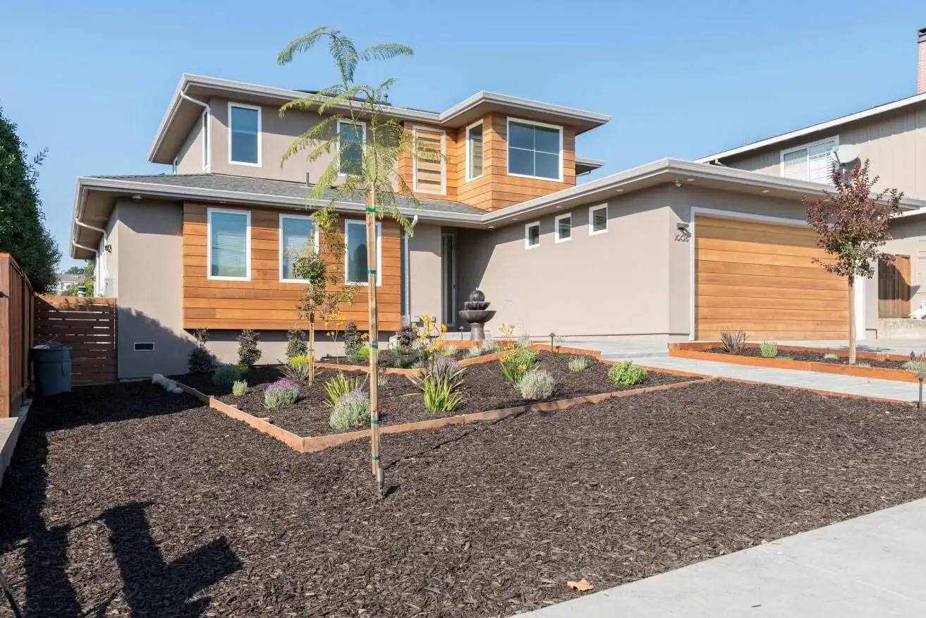 Two-story home exterior with wood-panel and stucco siding, a landscaped front yard with planting beds, and a wood-tone garage door
