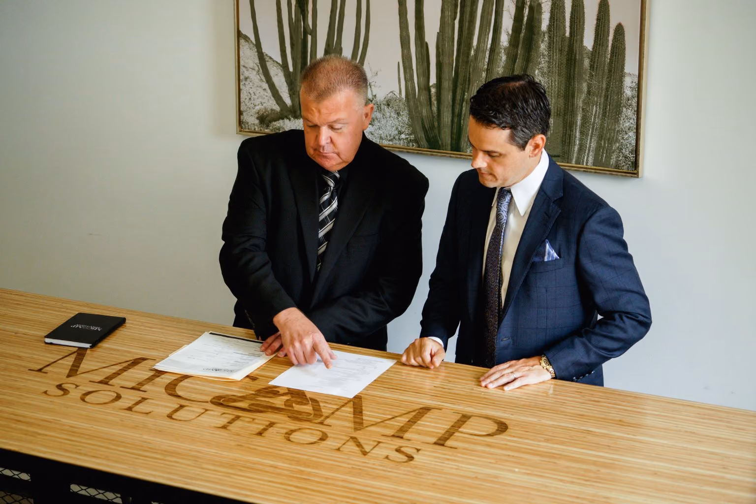 Two men in suits discussing documents on a wooden table with MicAmp Solutions logo.