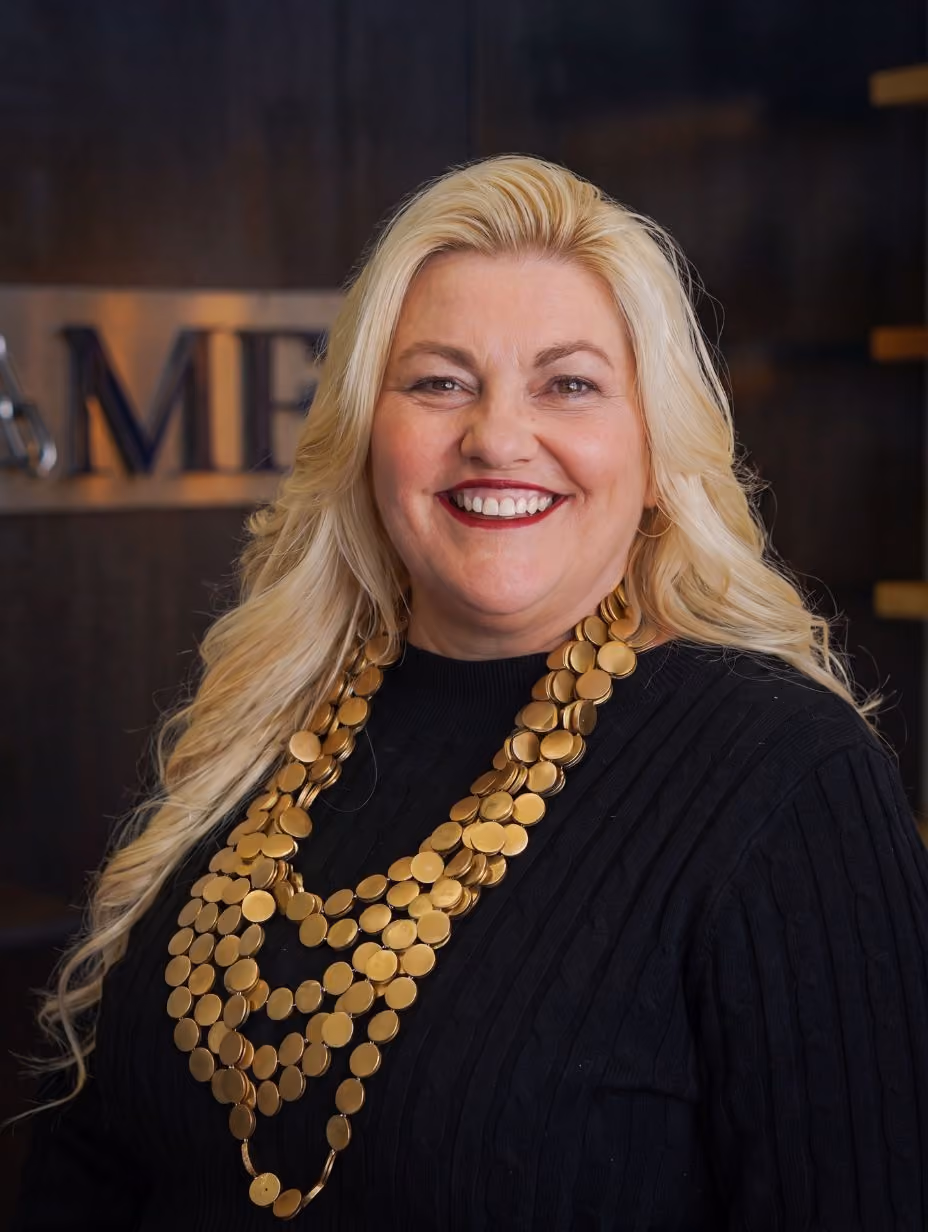Smiling blonde woman with long hair wearing a black top and a large gold coin necklace.