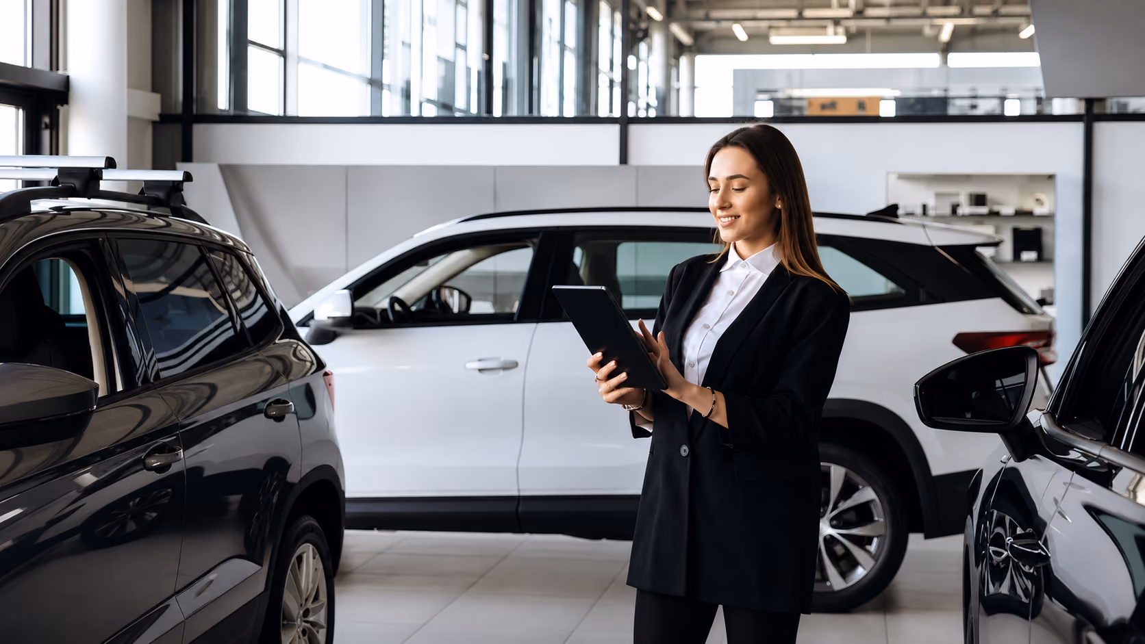 Woman in black blazer using a tablet inside a modern car dealership with white and black SUVs.
