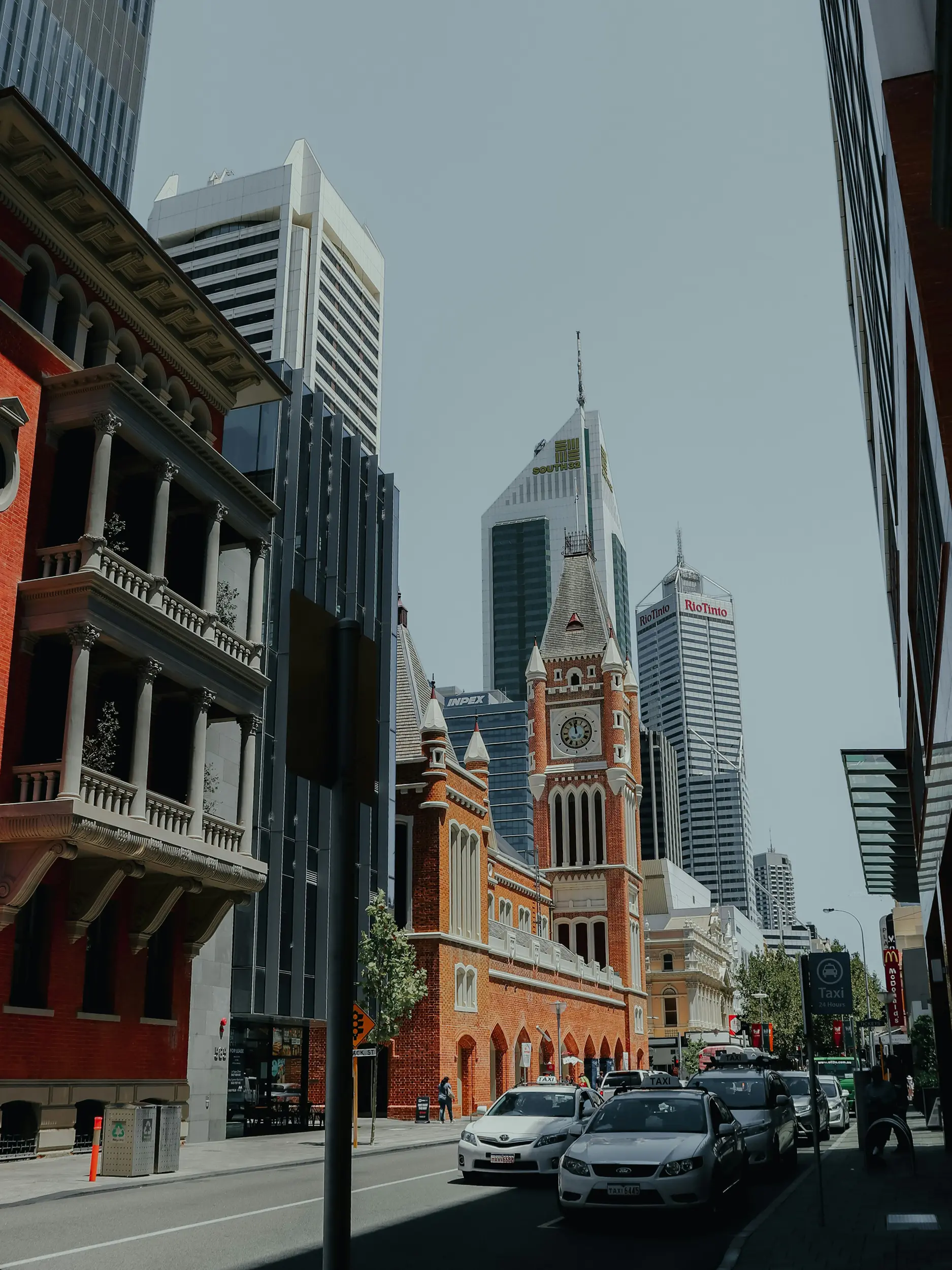 Street view of historic red brick clock tower building with taxis and modern skyscrapers in background.