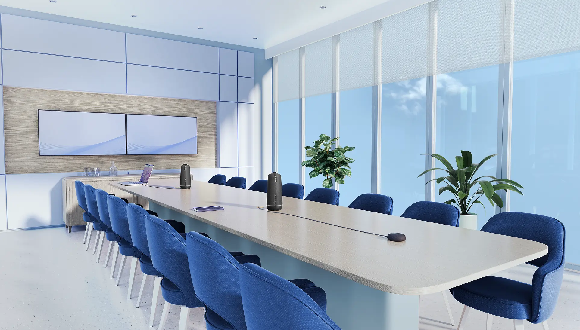 Modern conference room with a long rectangular light wood table surrounded by sixteen blue cushioned chairs, two black speakerphones on the table, a laptop, notebooks, and two large wall-mounted flat screens. The room has large floor-to-ceiling windows with sheer roller shades, allowing blue sky and clouds to be visible. There are two potted green plants near the windows and built-in cabinetry along the back wall.