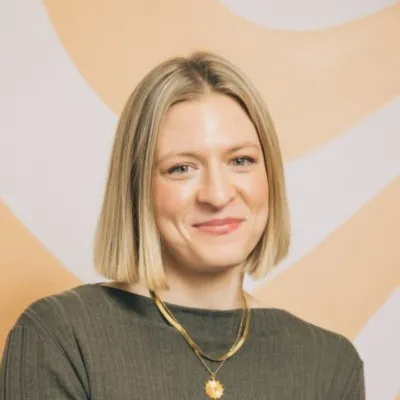 Portrait of a woman with shoulder-length blonde hair wearing a dark green top and gold necklace, smiling gently against a blurred beige and white abstract background.