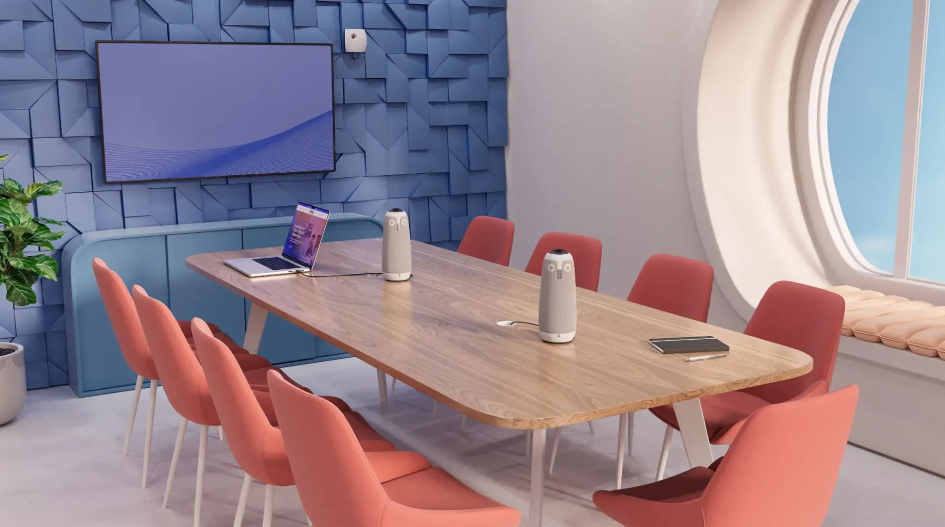Modern conference room with a light wooden table surrounded by eight coral upholstered chairs, two smart speakers on the table, an open laptop, a notebook with a pen, blue textured wall with a flat screen, a large round window with cushion seating, and a potted plant.