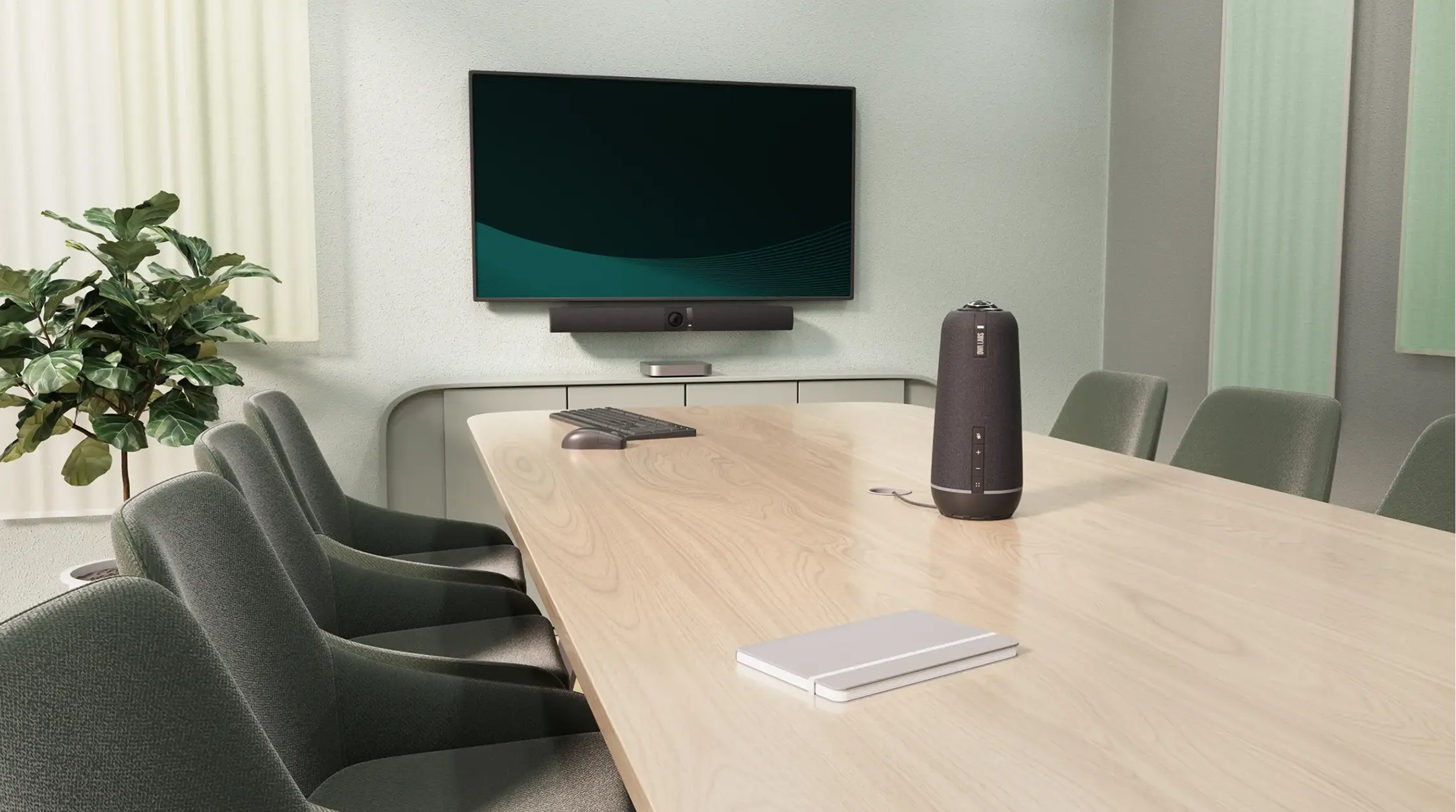 Modern conference room with a light wooden table, six green upholstered chairs, a large flat-screen TV mounted on the wall, a soundbar speaker beneath the TV, a wireless keyboard and mouse on the table, a closed notebook, a black cylindrical speakerphone device at the center, and a green potted plant in the corner.