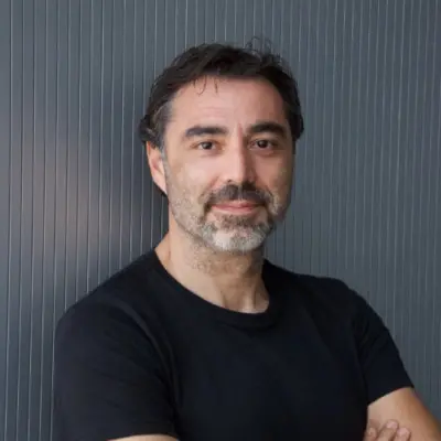 Man with dark hair and beard wearing a black t-shirt, standing with arms crossed in front of a vertical striped gray wall.