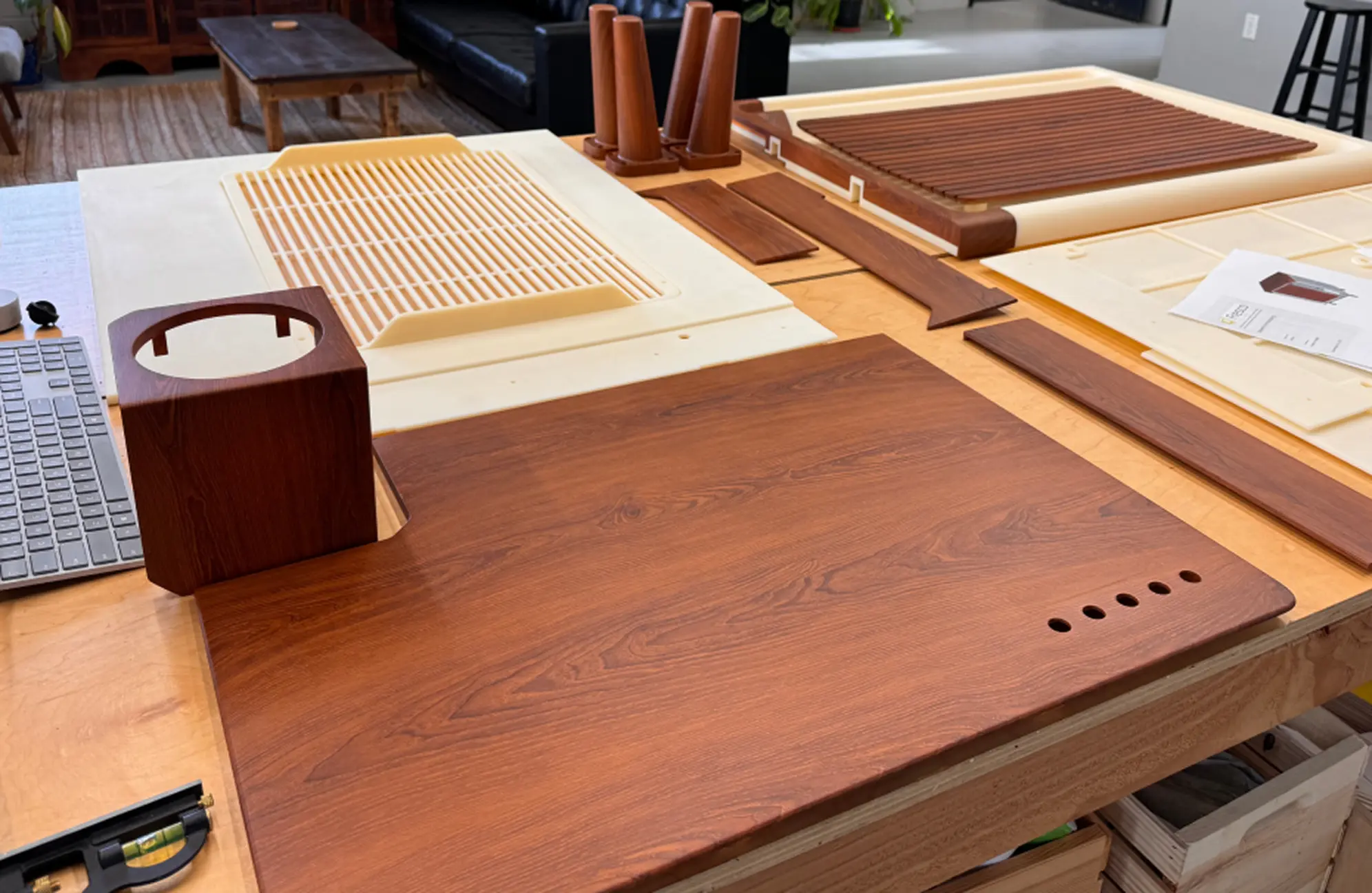 Various wooden pieces and components arranged on a workshop table, including a large flat panel with six round holes, a cube-shaped part with a circular cutout on top, and several wooden slats and rods, with a keyboard visible on the left side.