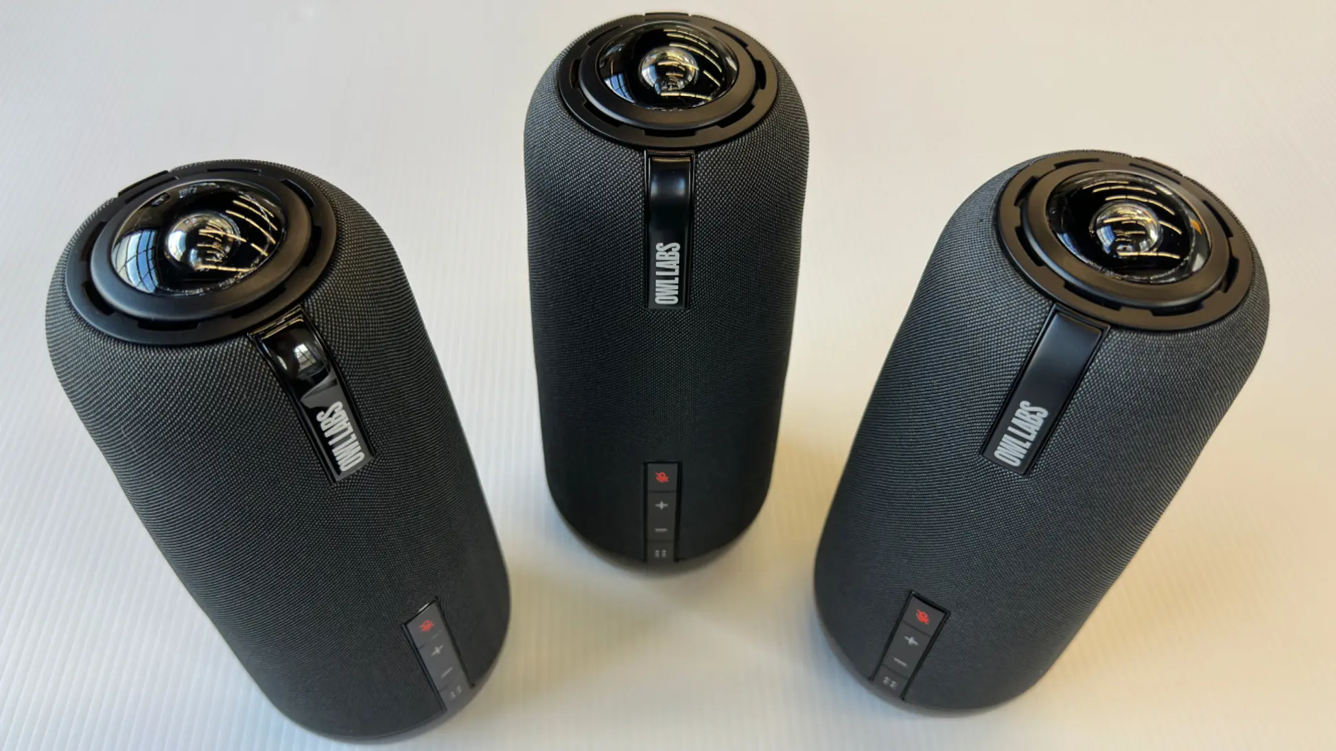Three black cylindrical Owl Labs smart 360-degree video conferencing cameras with fabric covers and lens domes on top, placed on a white surface.