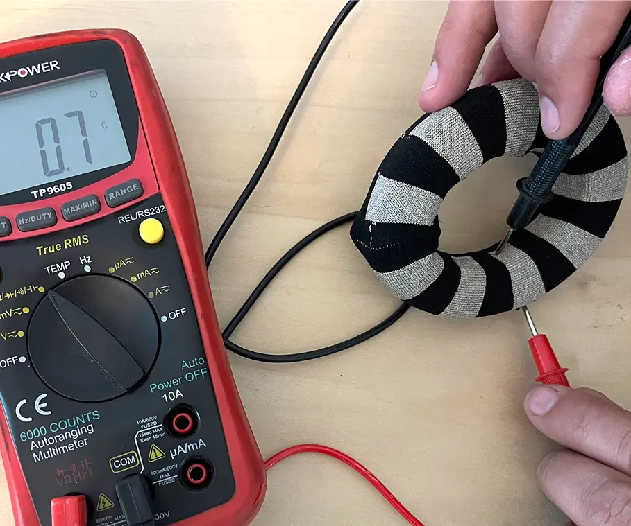 Hands using a red and black digital multimeter to measure resistance on a black and beige striped fabric ring, displaying 0.7 ohms on the screen.