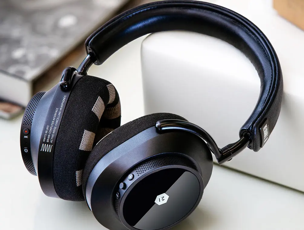 Black over-ear headphones with padded headband and ear cups resting on a white surface, featuring buttons and text indicating active noise cancelling and brain-computer interface.