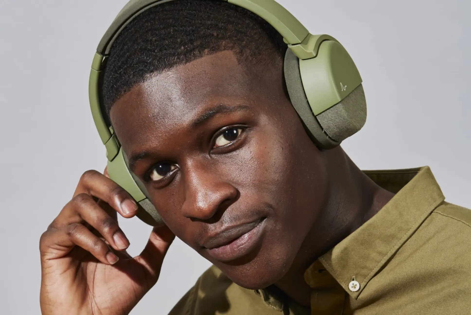 Close-up of a young man wearing olive green over-ear headphones and an olive green collared shirt, with one hand adjusting the headphones.