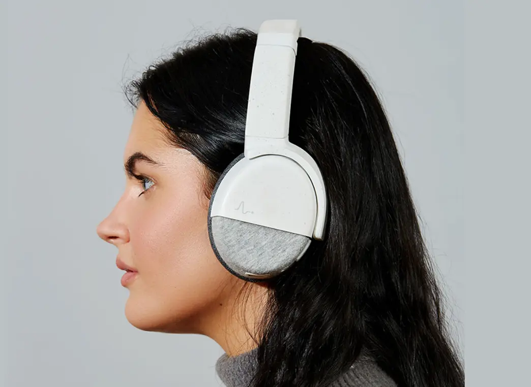 Woman with long black hair wearing large white over-ear headphones against a plain gray background, shown in profile view.