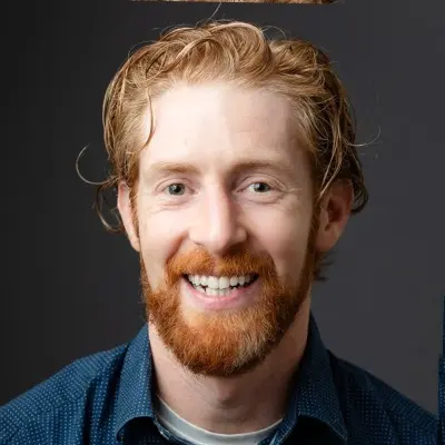 Portrait of a smiling man with red hair, a red beard, and wearing a blue button-up shirt against a dark background.