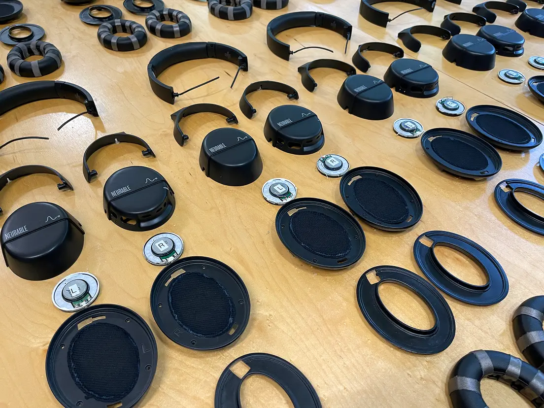Disassembled black over-ear headphones parts laid out on a wooden surface, including ear cushions, headbands, speaker drivers labeled L and R, outer casings with 'Neurable' branding, and internal components.