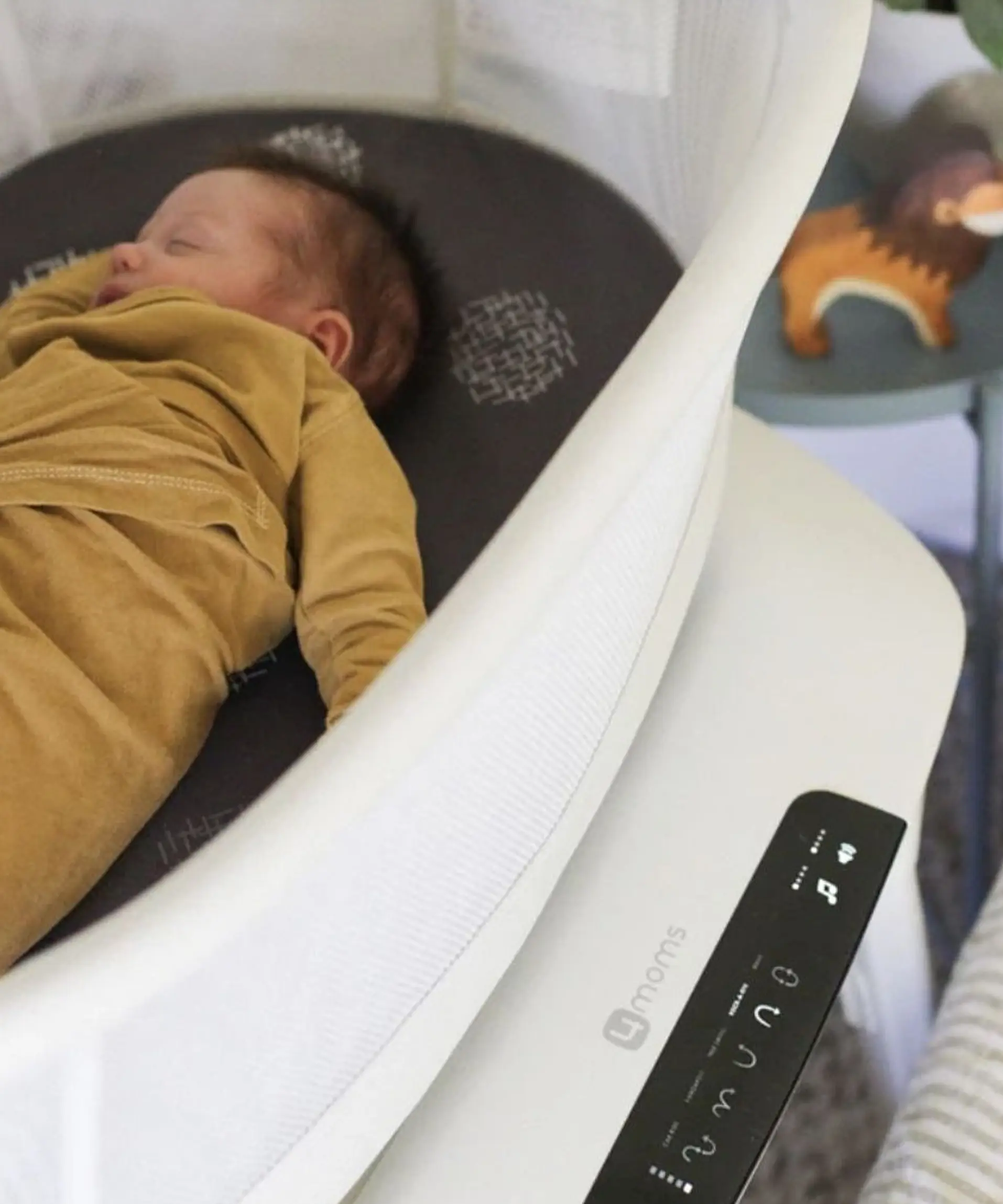 Sleeping baby dressed in a brown outfit lying in a white 4moms bassinet with a visible control panel, and a blurred lion-shaped toy in the background.