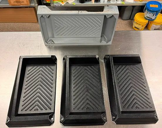 Four rectangular plastic trays with angled grooves arranged side by side on a metal surface, three black trays at the bottom and one gray tray at the top, with shelves and yellow and blue paint buckets in the background.