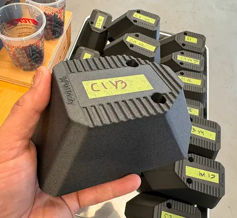A hand holding a black, textured plastic component with a yellow label reading 'C1 V3'; more similar components with yellow labels are stacked in the background on a gray surface.
