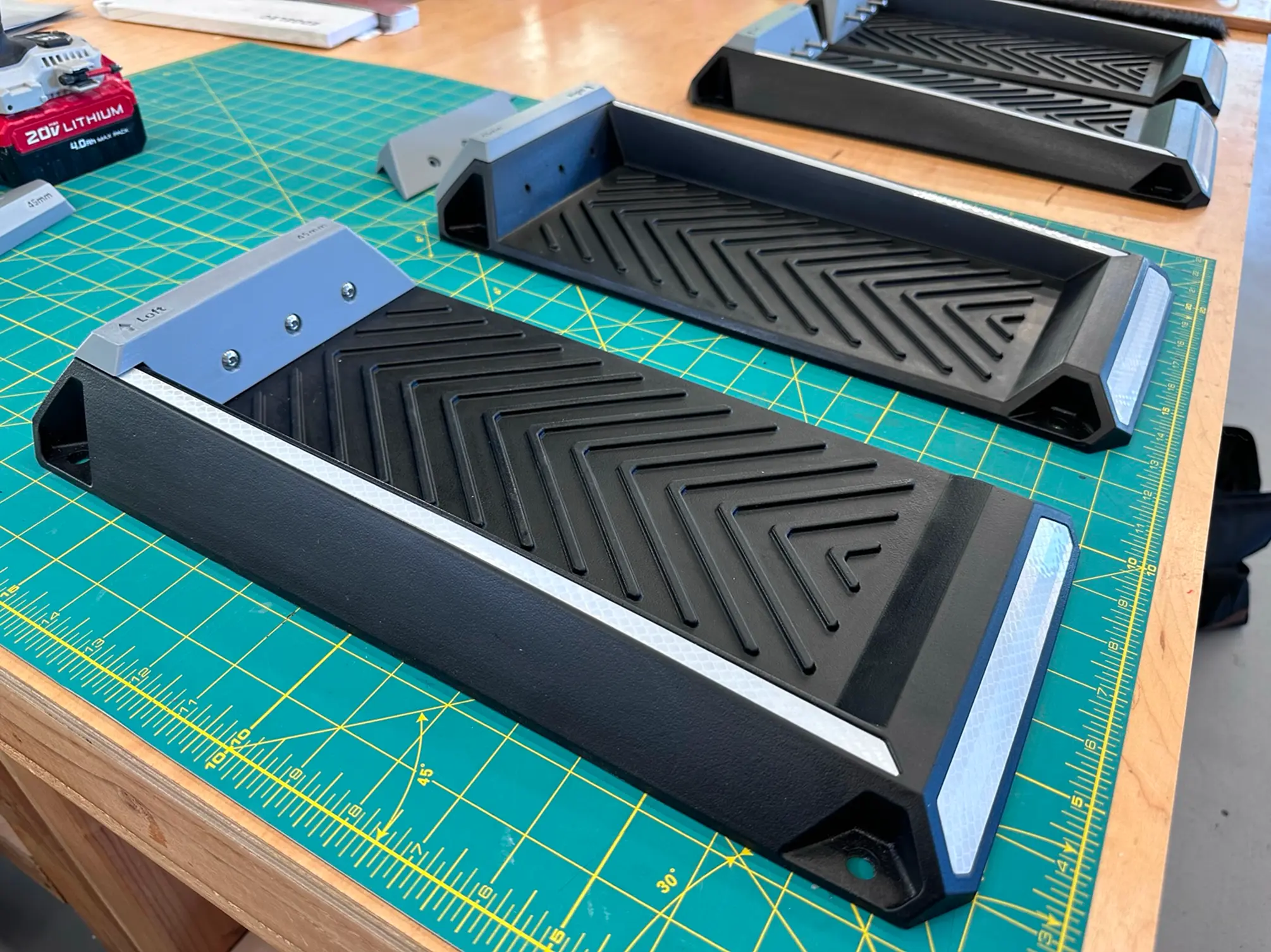 Black plastic pedal pads with diagonal ridges and reflective strips at the ends, arranged on a green cutting mat on a wooden workbench with a 20V lithium battery in the background.